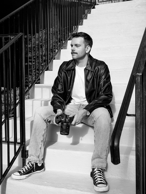 Black and white photo of Jeremy Uni sitting on steps holding camera